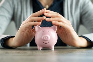 Close up of hands of woman protecting piggy bank for saving. Hands covering savings on table. Closeup of pink piggy bank protected by woman hands. Save money, investment and financial safety concept.