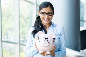 Closeup portrait happy smiling business woman holding pink piggy bank wearing big black glasses isolated indoors office background. Financial budget savings smart investment concept